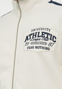 Λευκό μπουφάν με φερμουάρ και μπλε σκούρο κεντημένο έμβλημα στο στήθος που αναγράφει "University Athletic League Club 1987 Established Fear Nothing".