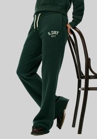 Pantalons de survêtement verts avec une coupe décontractée, arborant un logo blanc "S.DRY N.Y.C." sur la cuisse gauche et des cordons de serrage à la taille. Design à l'avant plat.