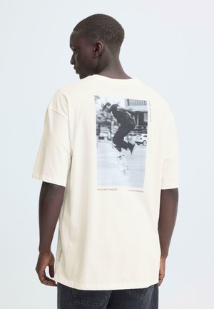 Kurzärmeliges Creme-T-Shirt mit einer schwarz-weißen Skateboard-Grafik auf der Rückseite und dem Text "SKATE WITH PURPOSE", lockere Passform.