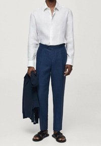 Witte linnen lange mouwen shirt, donkerblauwe op maat gemaakte broek, zwarte sandalen, houdt een marineblauwe jas vast. Soepele textuur en gestructureerde pasvorm.