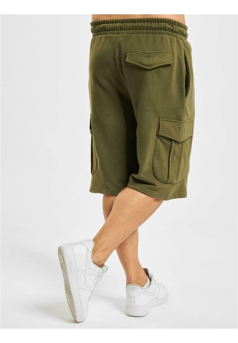 Personne portant un short cargo vert olive avec des poches à rabat, jambes croisées, et des baskets blanches, debout sur un fond blanc uni.