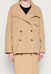 Beżowa podwójnie zapinana marynarka z szerokim kołnierzem, dużymi guzikami i luźnym krojem. Materiał wydaje się gładki i lekki.