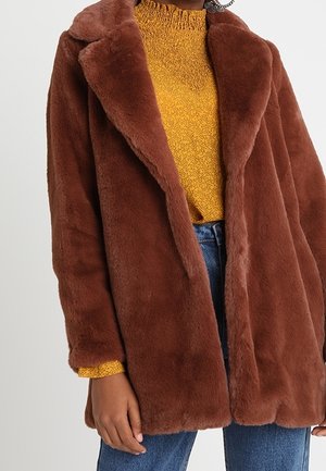 Personne portant un manteau en fausse fourrure marron par-dessus un pull texturé jaune et un jean bleu sur un fond uni.