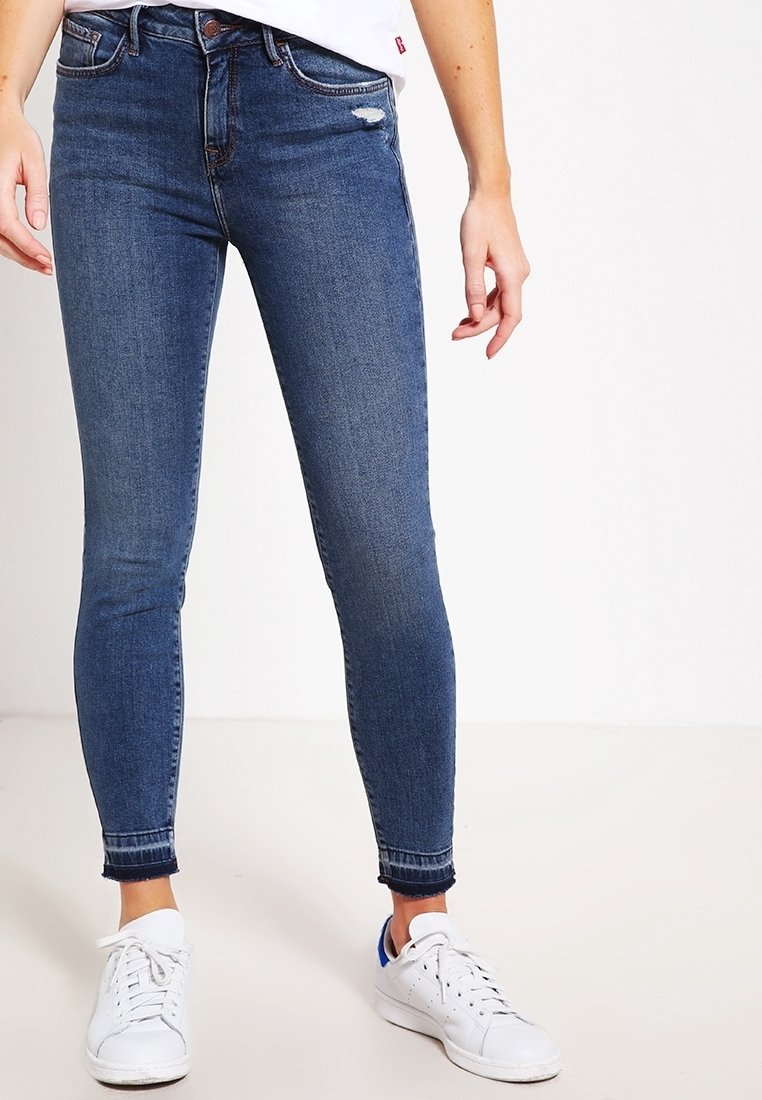 Jean skinny bleu avec un léger détail déchiré, fabriqué en denim, avec une taille mi-haute et des coutures contrastantes, associé à des baskets blanches.
