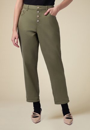 Pantaloni verde oliva a vita alta con tasche anteriori, caratterizzati da cinque bottoni decorativi. Orlo risvoltato sopra la caviglia con scarpe ballerine rosa a punta.