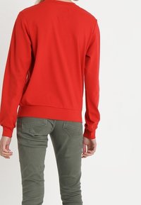 Person in einem leuchtend roten Langarmshirt und olivgrünen Hosen, steht mit dem Rücken zur Zuschauerschaft.