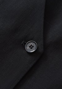 Schwarzer Stoff mit einer subtilen Textur, der an der Kante eines Kleidungsstücks einen runden, dunklen Knopf mit zwei Löchern aufgenäht hat.