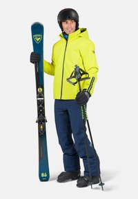 Equipo de esquí que incluye una chaqueta amarilla vibrante, pantalones azul marino, guantes negros, esquís azules con detalles verdes, bastones de esqu�í y un casco negro.