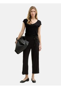 Top nero con maniche corte e scollatura a V, abbinato a pantaloni neri a gamba larga. Tiene in mano una grande borsa nera strutturata. Le scarpe flat nere completano l'outfit.