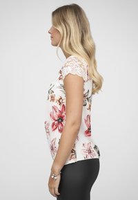 Bloes met bloemenmotief en korte kanten mouwen, met een witte achtergrond en grote rode en roze bloemen. De stof oogt glad en licht van gewicht.