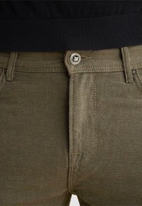 Gros plan d’un pantalon texturé kaki avec un bouton en métal orné d’un emblème de lion et des passants de ceinture, associé à une ceinture noire côtelée.