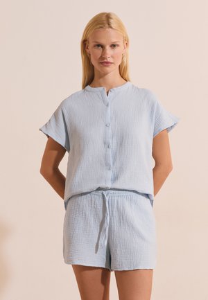 Blonde woman wearing light blue short-sleeve button-up top and matching drawstring shorts, standing with hands behind back.