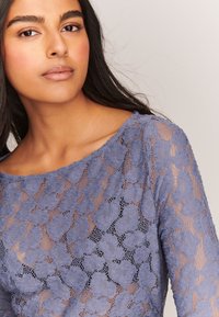 Top in pizzo di un delicato azzurro con motivo floreale, maniche lunghe e scollatura ampia, con texture trasparente e dettagli raffinati.