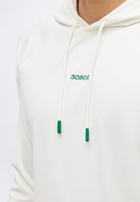 Λευκό φούτερ με κουκούλα με πράσινη κεντημένη επιγραφή "30904" στο στήθος και λευκά κορδόνια με πράσινες άκρες, φορεμένο από ένα άτομο.