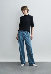 Γυναίκα που στέκεται φορώντας μαύρο κοντομάνικο ριμπ τοπ, φαρδιά μπλε τζιν με μικρές λευκές βούλες και λευκά μυτερά παπούτσια, μπροστά σε απλό τοίχο.