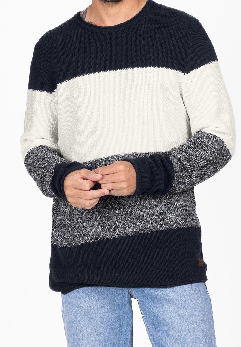 Homme portant un pull en tricot avec des blocs de couleur horizontaux bleu marine, blanc et gris et un jean bleu clair, mains jointes devant.