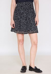 Kvinna klädd i en knälång svart kjol med mörka leopardprickar och svarta franslöpare, stående mot en enkel bakgrund.