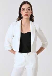 Blazer court blanc à manches trois-quarts, associé à un top noir et un pantalon blanc assorti, taillé sur mesure. Tissu lisse, design structuré.
