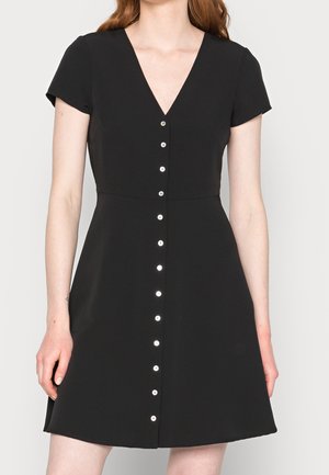 Kobieta ubrana w czarną sukienkę z krótkim rękawem, dekoltem w serek i przednim rzędem białych guzików, stojąca na jasnym, jednolitym tle.