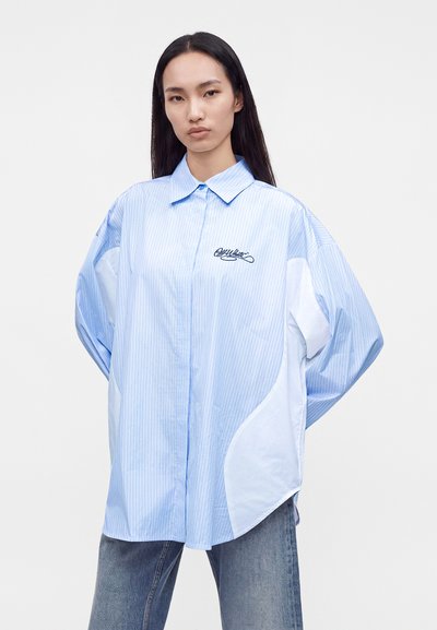 Femme aux longs cheveux foncés porte une chemise oversize bleu clair à fines rayures avec un panneau blanc et un logo brodé foncé sur la poitrine.
