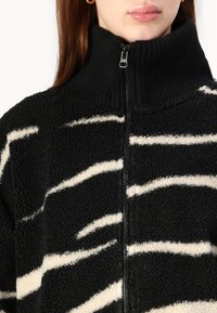 Svart fleecejakke med hvite zigzag-striper, høy ribbestrikket krage og glidelåsdetalj foran. Myk tekstur og uformell design.