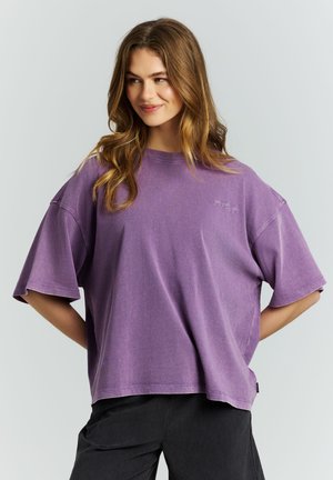 Jonge vrouw met lang haar, gekleed in een losse paarse T-shirt en zwarte broek, staand met haar handen in de zij tegen een neutrale achtergrond.