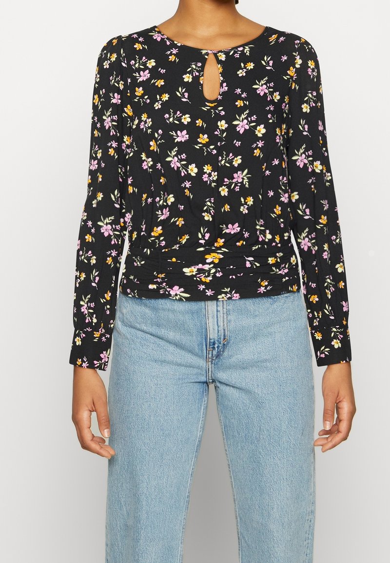 Svart blommig långärmad topp med nyckelhålshalsringning, prydd med rosa och gula blommor, samlad midja och mjuk tygtextur. Kombinerad med ljusblå jeans.