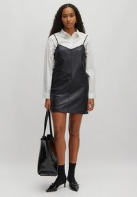Robe mini en cuir noir avec des bretelles spaghetti, associée à une chemise blanche à boutons, des chaussettes noires, des talons pointus et un sac fourre-tout noir.