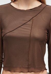 Καφέ μακρυμάνικο τοπ από μαλακό, διάφανο ύφασμα με στρογγυλή λαιμόκοψη και ασύμμετρη ραφή. Η ωμή ραφή στο τελείωμα προσθέτει υφή στο σχέδιο.