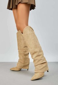Bottes en suédine beige à hauteur de genou avec un design flou, bouts pointus et un petit talon aiguille. Texture lisse avec des plis doux.