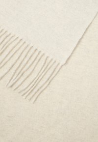 Myk kremfarget ullskjerf med frynser i endene liggende flatt på en matchende kremfarget stoffbakgrunn.