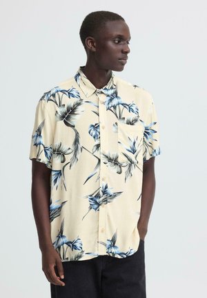 Ung mand iført en beige kortærmet skjorte med blå og sorte tropiske blomsterprint, kigger mod højre, hænderne i lommerne.
