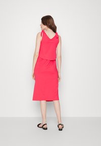 Vestido rosa sem mangas com alças em laço e saia solta, feito de um tecido suave, que repousa um pouco abaixo do joelho. Sandálias pretas complementam o conjunto.