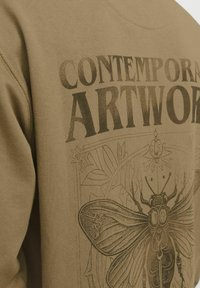 Sudadera color caqui con una gran impresión gráfica oscura de una mariposa y el texto "OBRA DE ARTE CONTEMPORÁNEA" en letras en negrita. Textura suave.