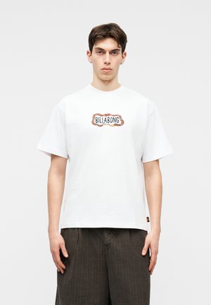 Young man wearing a white Billabong t-shirt with a patterned logo, paired with dark loose-fitting shorts, standing against a plain background.