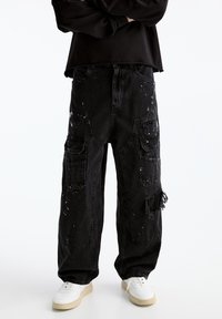 Pantalones cargo de mezclilla negros con un ajuste holgado, detalles desgastados, múltiples bolsillos y acentos de salpicaduras de pintura blanca.