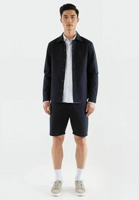 Marine strukturert skjorte med knapper, hvitstripet skjorte under, matchende marineblåshorts, hvite sokker og beige sneakers.