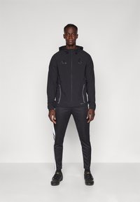 Giacca sportiva nera con zip e cappuccio, dotata di logo e due tasche laterali. Abbinata a pantaloni da tuta neri con strisce bianche.