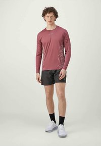 Langermet maroon sportskjorte med hvite grafiske mønstre på siden, parret med sorte shorts og hvite treningssko.