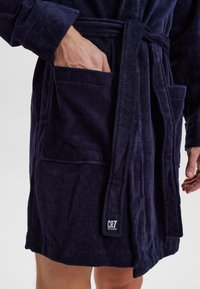 Person in einem dunkelblauen Frottee-Bademantel, mit einer Hand in der vorderen Tasche und einem gebundenen Gürtel mit einem CR7-Logo-Patch.