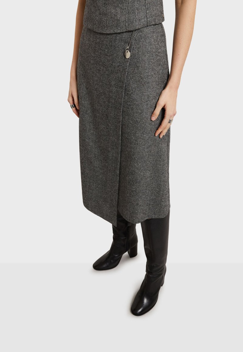 Siva suknja u riblju kost s oblikom omote, napravljena od teksturiranog materijala, uparena s crnim kožnim čizmama do bedrа. Suptilni detalj od metalnog hardvera.