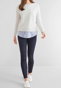 Sweat-shirt gris par-dessus une chemise boutonnée bleu clair, accompagné d'un jean bleu foncé et de baskets blanches. Tissu lisse, coupe décontractée, jambes fuselées.