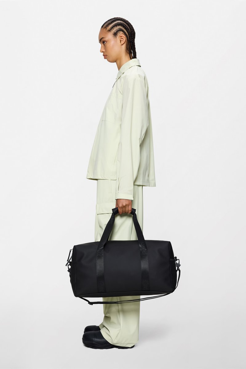 Μαύρη διπλή τσάντα duffel με διπλές υφασμάτινες λαβές, γυαλιστερή υφή και κλείσιμο με φερμουάρ, κρατημένη από άτομο με ανοιχτοπράσινη ενδυμασία σε απλό φόντο.