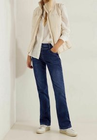 Chaleco acolchado beige con un cuello texturizado sobre una camisa blanca de botones, combinado con jeans rectos de pierna oscura y zapatillas blancas.