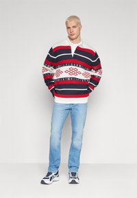 Suéter a rayas en colores azul marino, rojo y blanco con cuello, combinado con unos vaqueros azul claro y zapatillas deportivas.