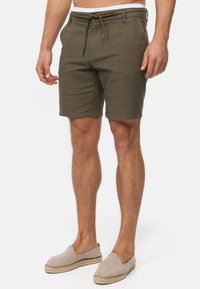Shorts vert olive avec une taille élastique à cordon de serrage, poches latérales et un tissu texturé. Portés avec des chaussures à enfiler beiges.