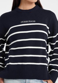 Pulover tricotat bleumarin cu dungi orizontale albe și "GUESS JEANS" brodat pe piept, purtat cu blugi deschiși la culoare.
