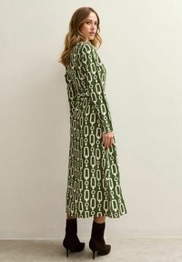 Vrouw die een olijfgroene midi-jurk met lange mouwen draagt, versierd met witte geometrische kettingpatronen, en zwarte enkellaarsjes met hakken, staand tegen een effen muur.