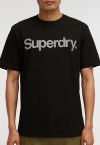 Camiseta de algodón negra con un gran logo reflectante en plata de "Superdry" en el pecho. Mangas cortas, diseño de cuello redondo estándar.