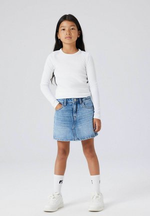 Mladá dívka stojí sebevědomě s rukama v kapse, má na sobě bílý dlouhý rukáv, džínovou sukni, bílé ponožky a bílé tenisky na jednoduchém pozadí.